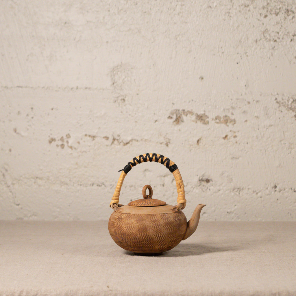 Brown ceramic teapot with a textured surface and wicker handle on a light gray background