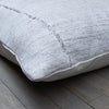 A close-up of a square throw pillow made from a textured, woven light gray linen fabric, resting on a rustic gray wooden floor.
