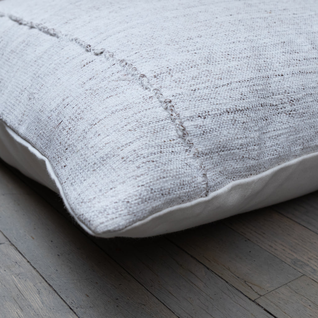 A close-up of a square throw pillow made from a textured, woven light gray linen fabric, resting on a rustic gray wooden floor.