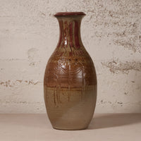 A handmade ceramic vase with a brown and beige gradient glaze, featuring a reddish-brown patterned neck and dripping glaze details, set against a textured off-white wall.