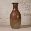 A handmade ceramic vase with a brown and beige gradient glaze, featuring a reddish-brown patterned neck and dripping glaze details, set against a textured off-white wall.