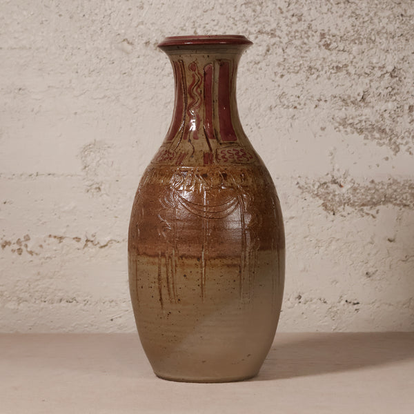 A handmade ceramic vase with a brown and beige gradient glaze, featuring a reddish-brown patterned neck and dripping glaze details, set against a textured off-white wall.