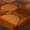 Close-up of a wooden table corner showing a mosaic tile top with circular and oval patterns in warm brown and tan tones, with a polished wood frame.