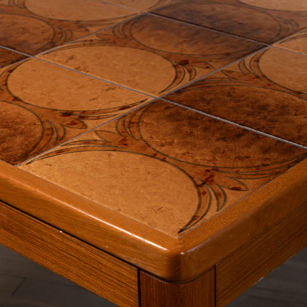 Close-up of a wooden table corner showing a mosaic tile top with circular and oval patterns in warm brown and tan tones, with a polished wood frame.