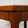 Close-up of a mid-century modern wooden table corner, showcasing a warm reddish-brown teak wood grain finish, drawer front, and tapered leg against a plain white background.