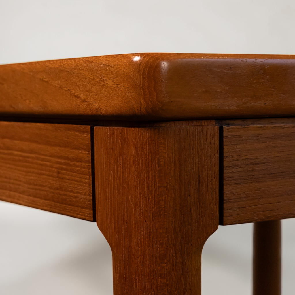 Close-up of a mid-century modern wooden table corner, showcasing a warm reddish-brown teak wood grain finish, drawer front, and tapered leg against a plain white background.