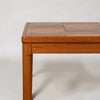 Close-up of a mid-century modern wooden table corner, showcasing a warm medium-brown wood grain top with a segmented geometric pattern and a matching solid wood leg, set against a plain light gray background.