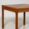 A close-up of a wooden table corner showing a warm medium-brown wood grain finish, square top with inset paneling, and straight legs on a light gray background.