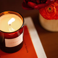 Candle with a visible label next to a vase with red flowers on a surface