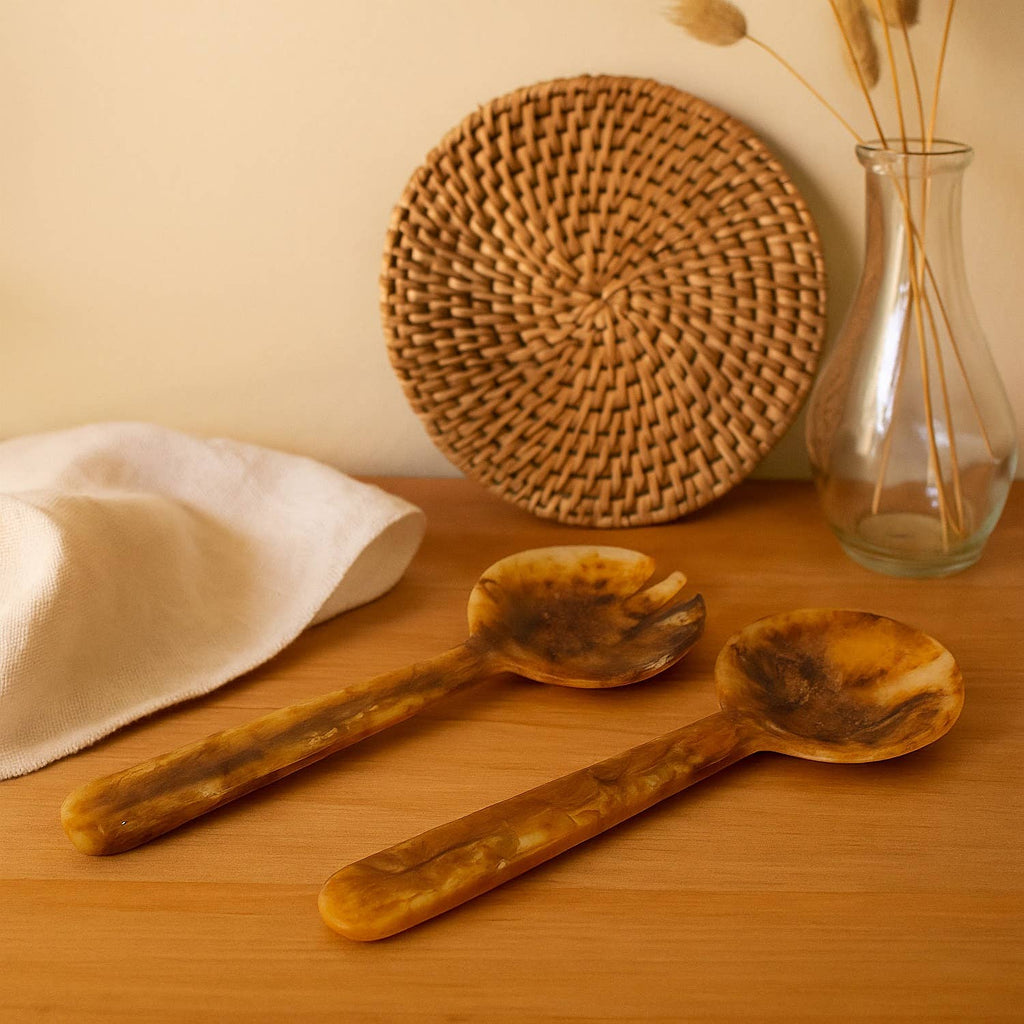 Two salad servers on a wooden surface with a woven placemat and vase in the background.