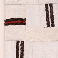A close-up of a woven textile or rug showing a patchwork-like pattern of off-white woven squares with dark brown stripes and one horizontal stripe featuring a red accent.