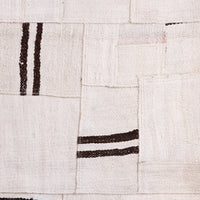 A close-up of a woven textile or rug made of patchwork-style rectangular panels in warm off-white/cream with dark brown striped accents.