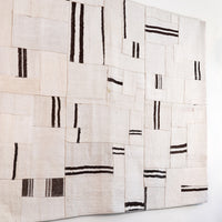 A square textile wall hanging or rug made from overlapping off-white woven fabric panels with irregular black stripe and dash patterns, shown against a plain white wall.