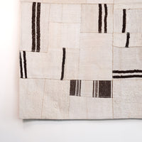 A close-up of a woven textile or rug with a patchwork design in warm off-white and beige, accented by dark brown and black vertical stripe patterns, shown against a plain white background.
