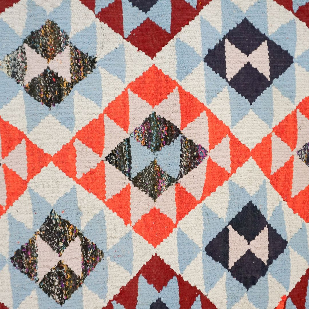 A close-up of a woven textile with a geometric diamond and triangle pattern in light blue, coral orange, cream, dark navy, and burgundy, with some multicolored textured centers.