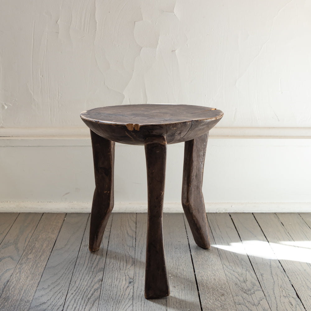 A rustic dark wood stool with a round seat and three tapered legs, set on a gray wooden plank floor against a white textured wall.
