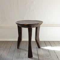 A rustic dark wood stool with a round seat and three tapered legs, set on a gray wooden plank floor against a white textured wall.