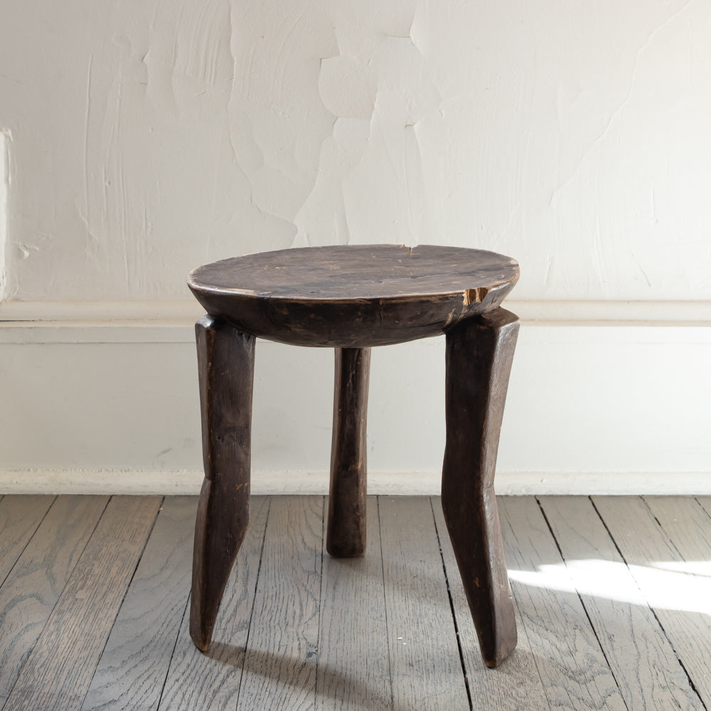 A rustic dark brown carved wooden stool with a round seat and three twisted legs, set on a weathered wood floor against a textured off-white wall.