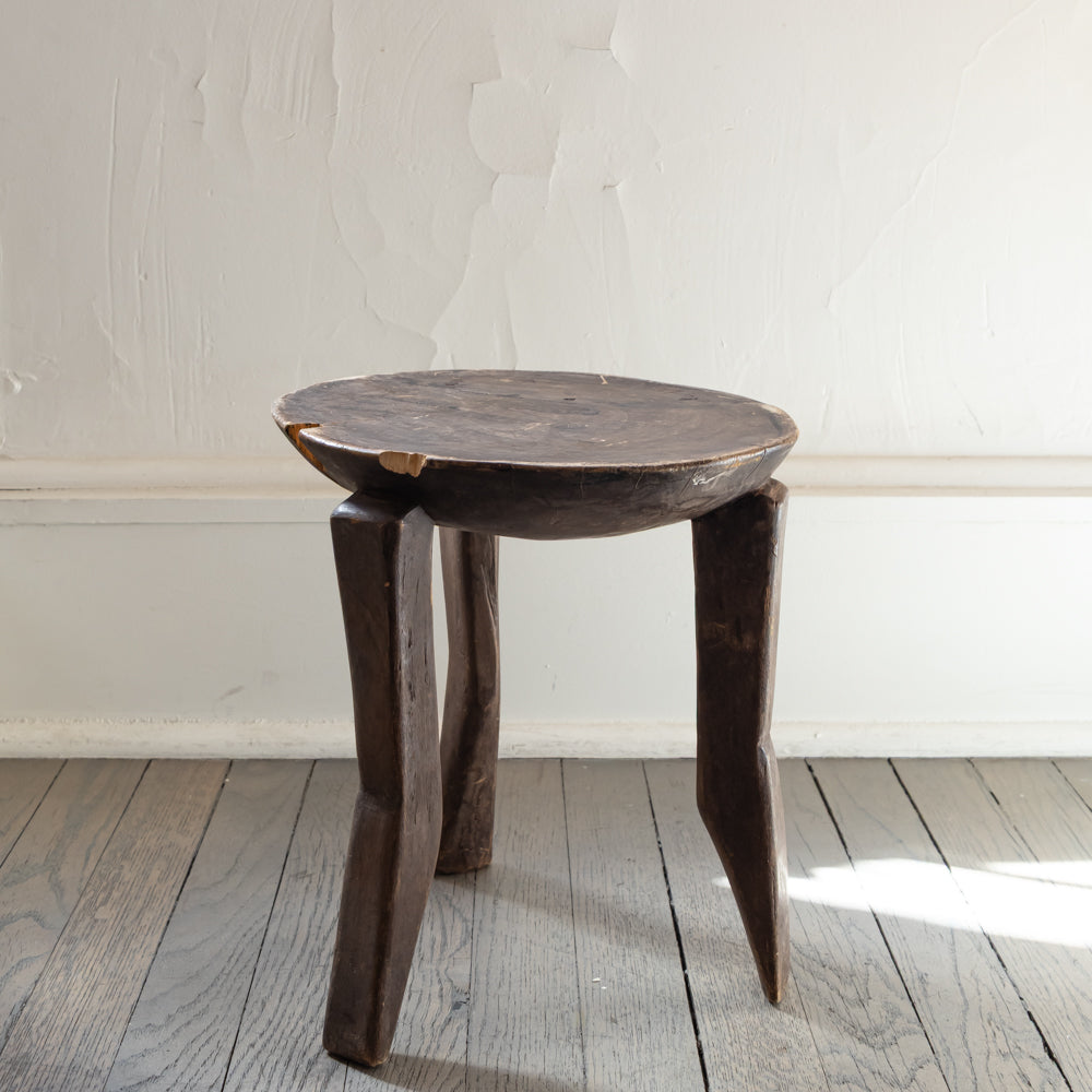 A rustic dark brown carved wooden stool with a round seat and three tapered legs, set on a gray wooden plank floor against a white textured wall.
