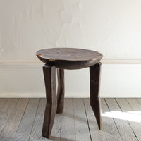 A rustic dark brown carved wooden stool with a round seat and three tapered legs, set on a gray wooden plank floor against a white textured wall.