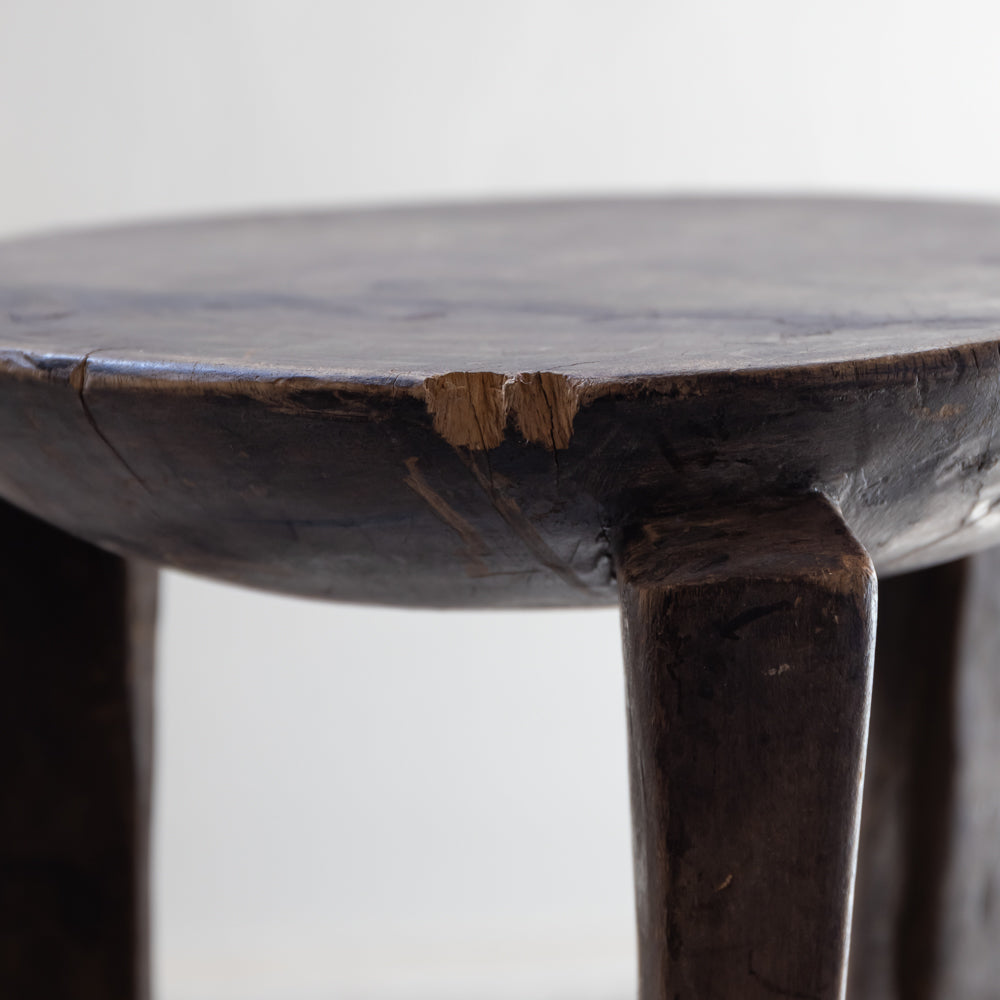 Close-up of a dark wooden stool showing the top edge of the seat with a visible crack and worn wood showing through, supported by thick legs against a plain light background.