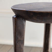 Close-up of a dark rustic wooden stool showing the thick round seat and one of its sturdy legs, set against a softly blurred light background.