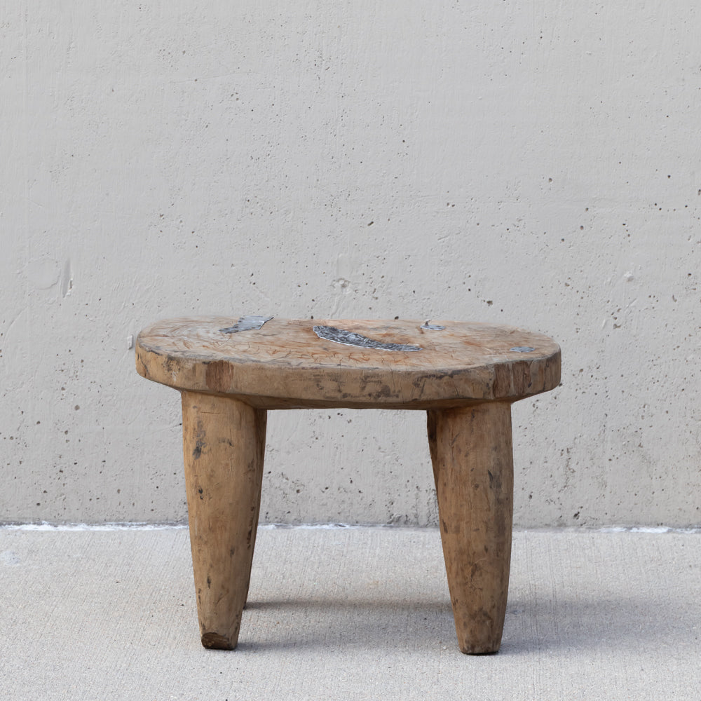 A rustic, low-profile wooden stool made from a thick, round natural wood slab with three sturdy legs, set against a textured concrete wall and floor.