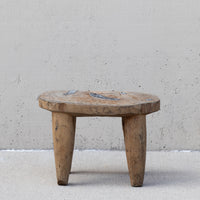 A rustic, low-profile wooden stool made from a thick, round natural wood slab with three sturdy legs, set against a textured concrete wall and floor.