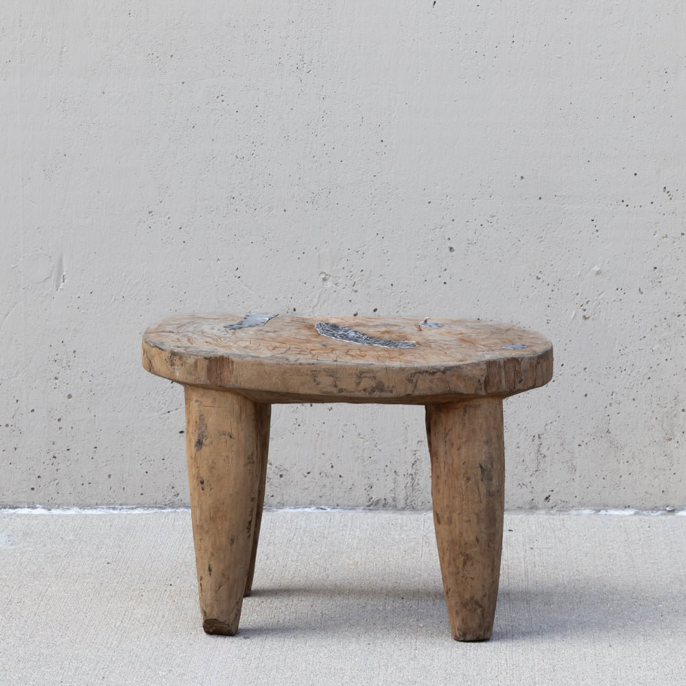 A rustic, low-profile wooden stool with a round seat and four tapered legs, positioned against a textured concrete wall.