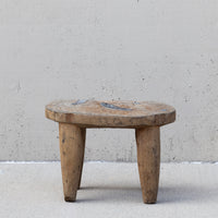 A rustic, low-profile wooden stool with a round seat and four tapered legs, positioned against a textured concrete wall.