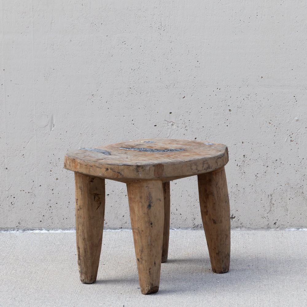 A rustic three-legged wooden stool made from a natural tree trunk, set against a textured concrete wall and floor.