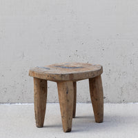 A rustic three-legged wooden stool made from a natural tree trunk, set against a textured concrete wall and floor.