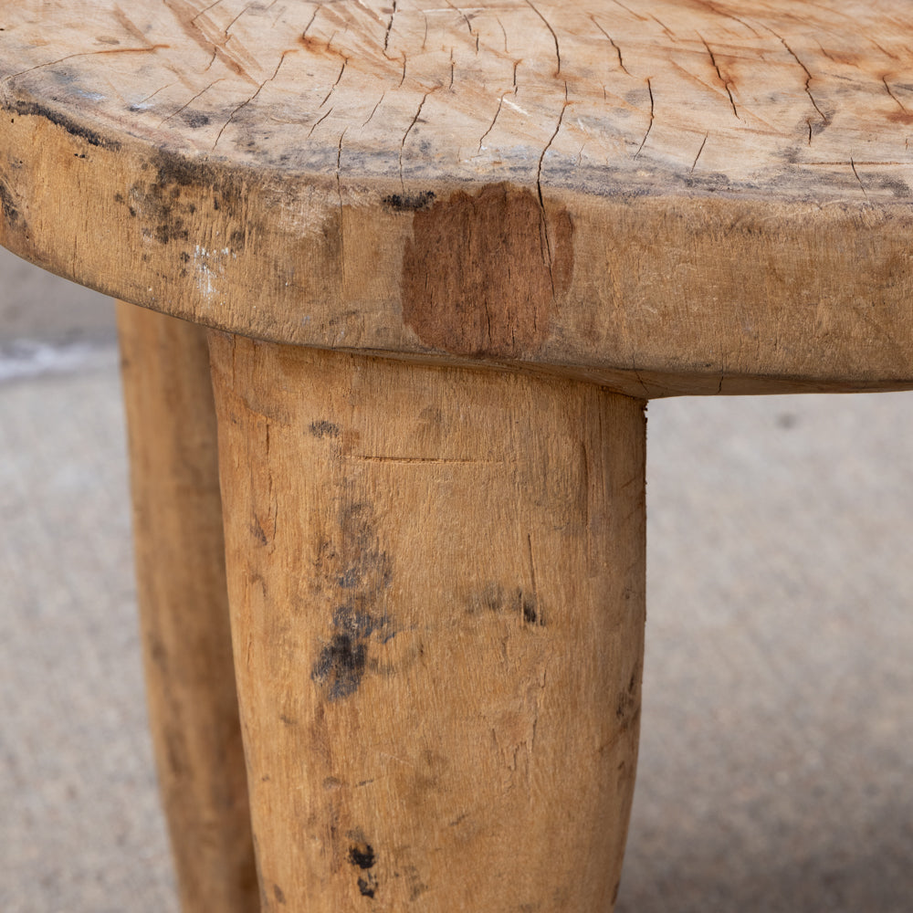 Close-up of a rustic wooden stool with a thick, round log-top surface and thick, rounded legs showing natural wood grain, knots, and weathered marks.
