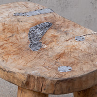 A rustic wooden stool with a thick, rounded natural wood slice top featuring visible grain, knots, and dark metal peg fasteners, shown in a close-up outdoor shot.