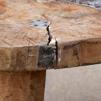 Close-up of a rustic wooden table with a thick natural wood slab top, visible grain, a prominent crack through the center, and dark metal hardware including a square bracket and round mounting plate.