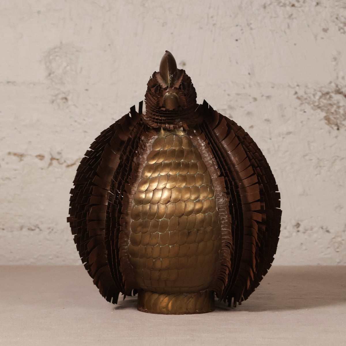 A decorative sculpture of a stylized bird or peacock made from metallic materials, featuring a golden-bronze scaled body and dark brown feather-like wings, set against a textured off-white wall.