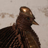 A close-up of a bird sculpture or decorative object made from layered metallic and wooden strips, with a polished copper-toned head and back and darker brown wings, set against a light textured wall.