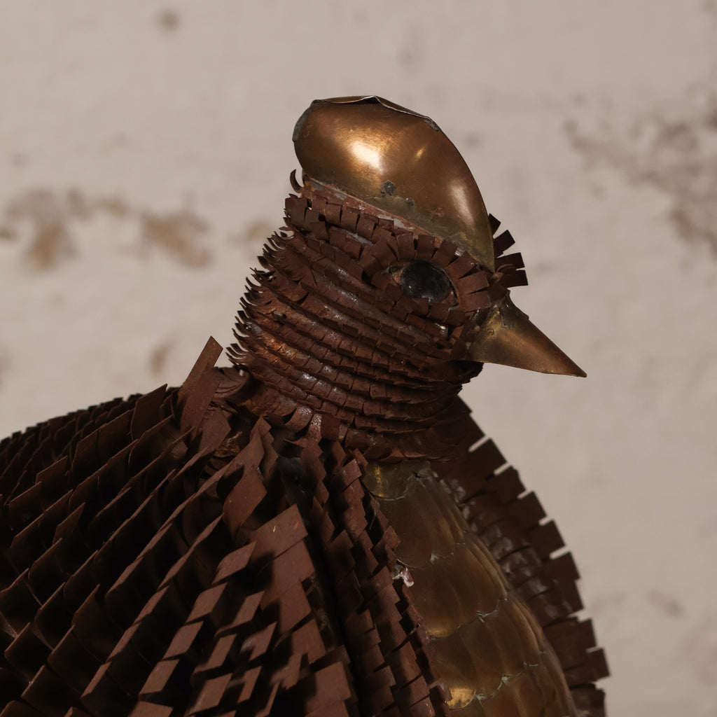 A close-up of a bird sculpture or decorative object made from layered metallic and wooden strips, with a polished copper-toned head and back and darker brown wings, set against a light textured wall.