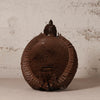 A dark brown woven decorative shell-shaped object with a textured, basket-like surface and a pointed top, photographed against a light off-white plaster wall.