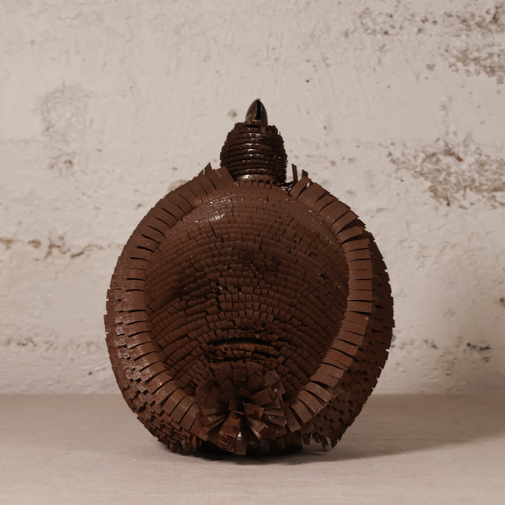 A dark brown woven decorative shell-shaped object with a textured, basket-like surface and a pointed top, photographed against a light off-white plaster wall.