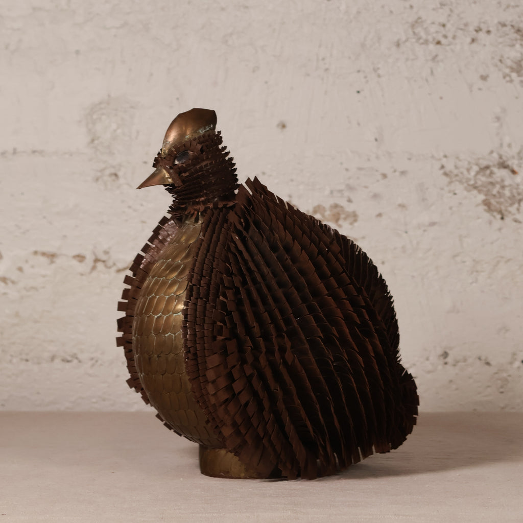 A sculptural bird figurine made from overlapping metallic scales, shown in profile against a textured off-white wall and tabletop.
