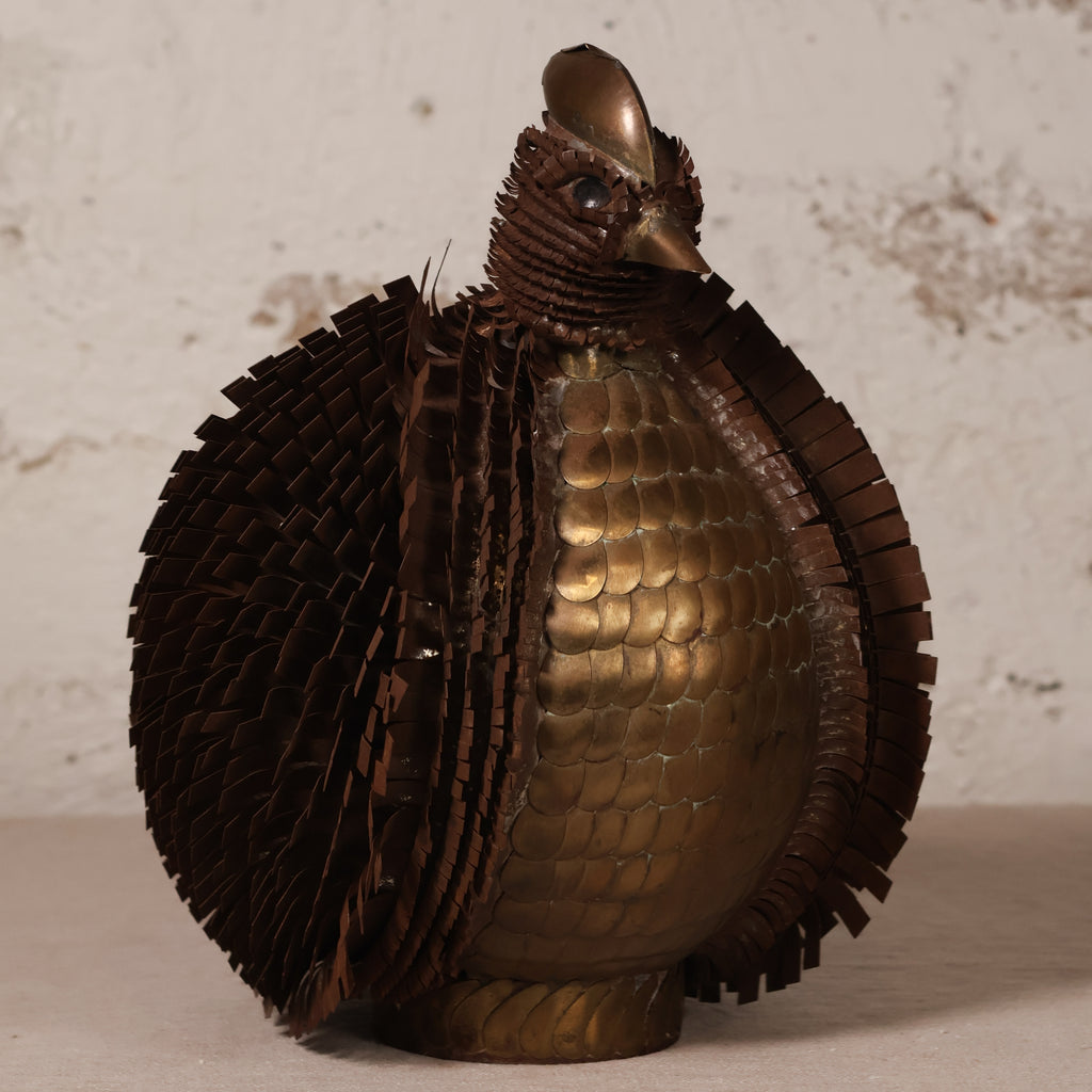 A decorative bird sculpture made from layered metallic materials, featuring a bronze-toned scaled body and dark brown feather-textured wings and neck, set against a light textured wall.