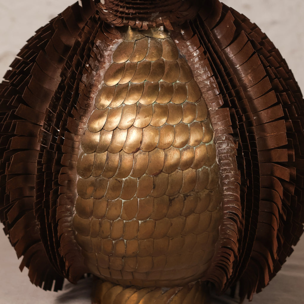 Close-up of a metallic bronze sculpture with a central rounded, scale-like textured section surrounded by layered, fringed, leaf-like extensions on a light neutral background.