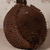 A dark brown woven decorative object made from layered ribbon or paper strips, photographed against a light neutral textured background.