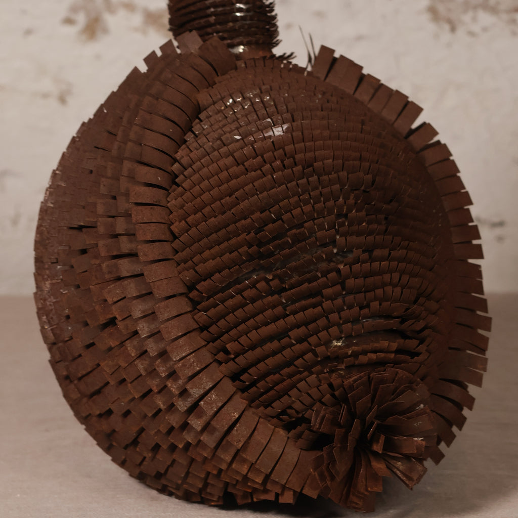 A dark brown woven decorative object made from layered ribbon or paper strips, photographed against a light neutral textured background.