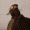 A close-up of a bird sculpture made from layered metallic copper and bronze sheets, showing the head and upper body against a plain light beige background.