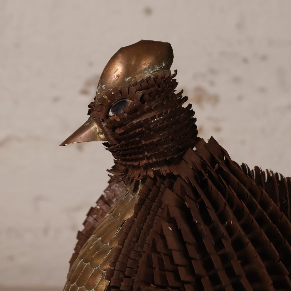 A close-up of a bird sculpture made from layered metallic copper and bronze sheets, showing the head and upper body against a plain light beige background.