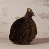 A sculptural bird figurine made from layered metallic strips, featuring a dark brown textured body and a polished bronze-toned head and beak, set against a rustic white plaster wall.