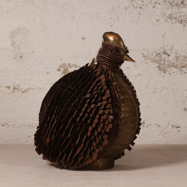 A sculptural bird figurine made from layered metallic strips, featuring a dark brown textured body and a polished bronze-toned head and beak, set against a rustic white plaster wall.