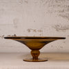 An amber glass pedestal plate with a wide, shallow bowl and a fluted stem, set against a textured, distressed off-white wall.